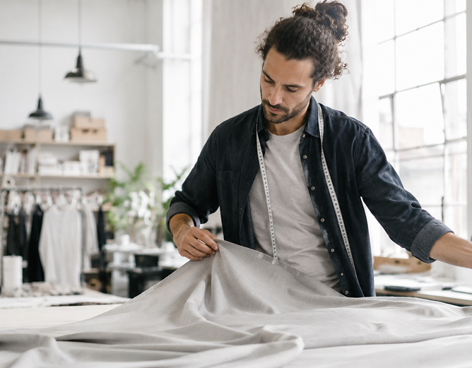 A person with a measuring tape spreads light-colored fabric across a large table in a bright workshop.
